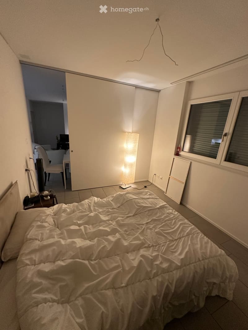 A furnished bedroom with a bed, nightstand, chair, table, lamp, and wardrobe. The room has tiled flooring and a window with blinds.