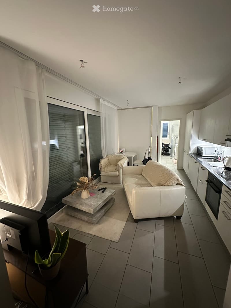 Modern living room with white sofa, armchair, coffee table, and a small table near the window. The room features tiled flooring, white walls, and a view of the kitchen area with cabinets and appliances.