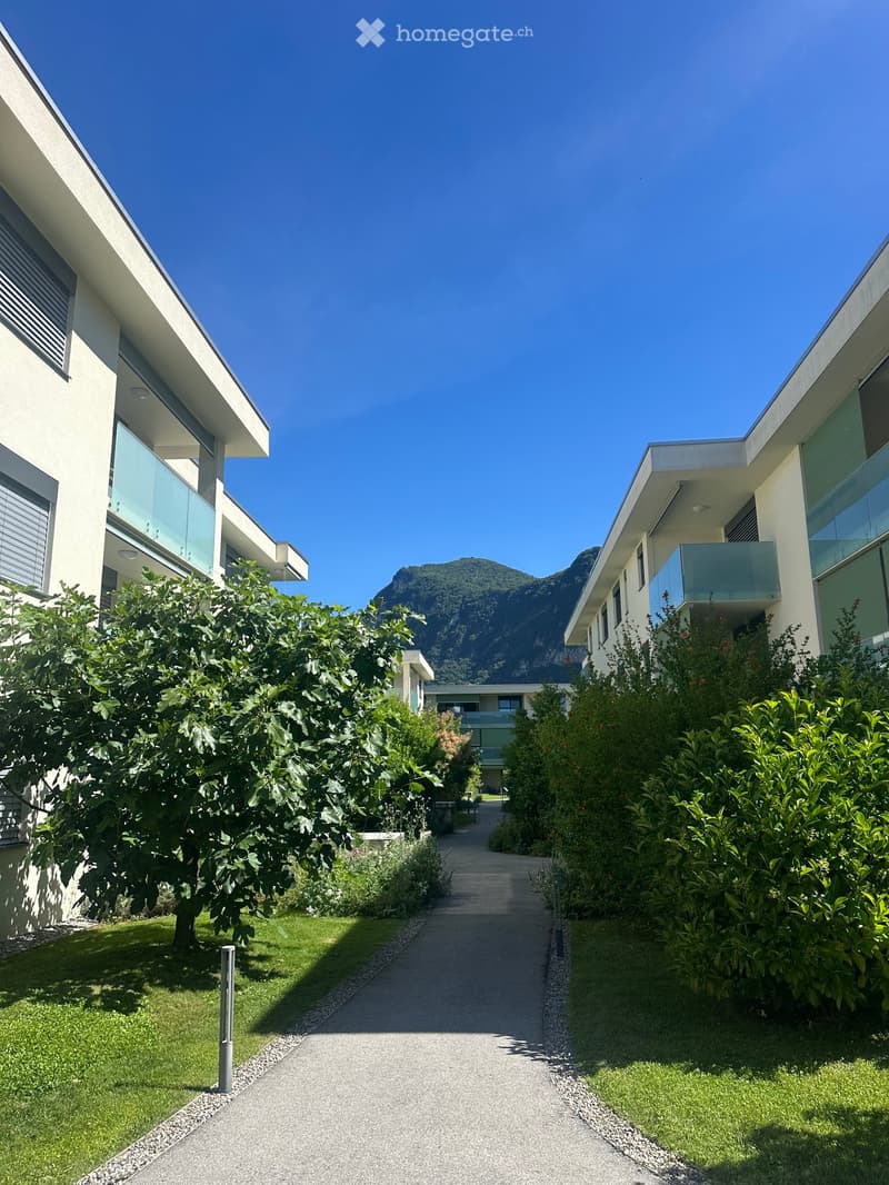 Modern residential complex with clean lines, glass balconies, and lush greenery, set against a backdrop of a mountain view.