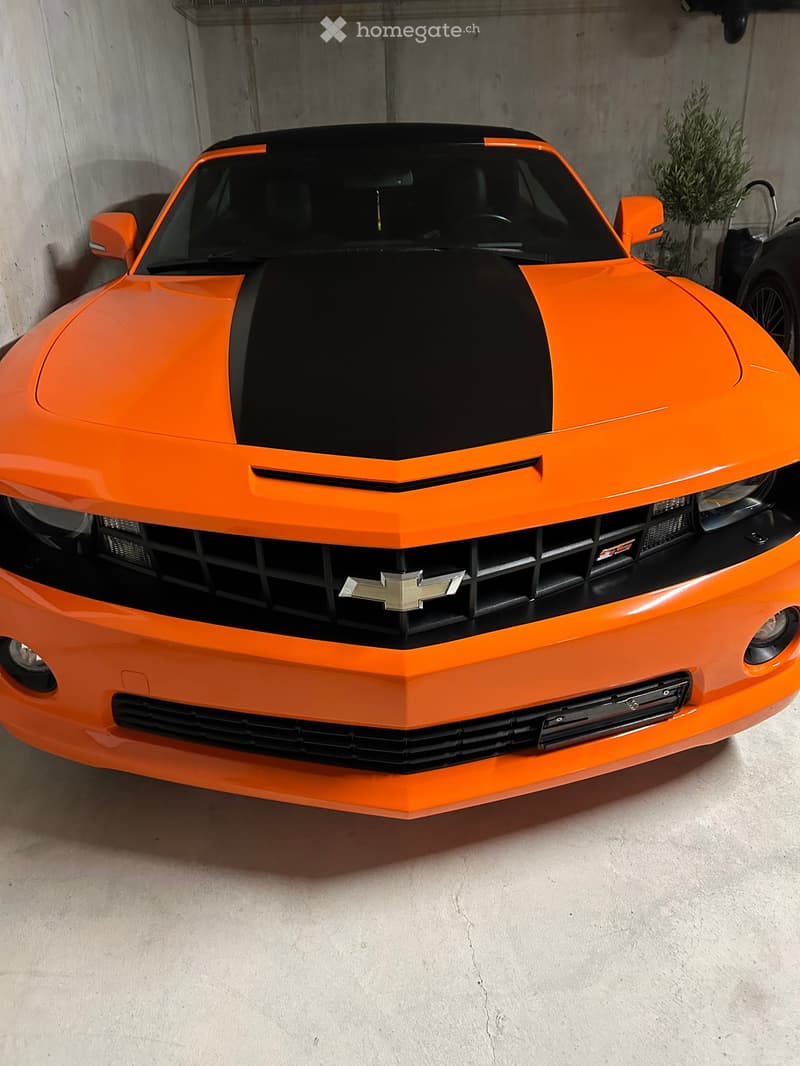 A vibrant orange sports car with a black hood and grille is parked inside a garage, with a small potted plant visible in the background.