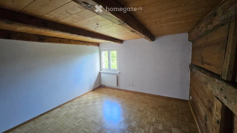 Empty room with wooden floor, exposed wooden beams on the ceiling, white walls, and a single window with a radiator below it.