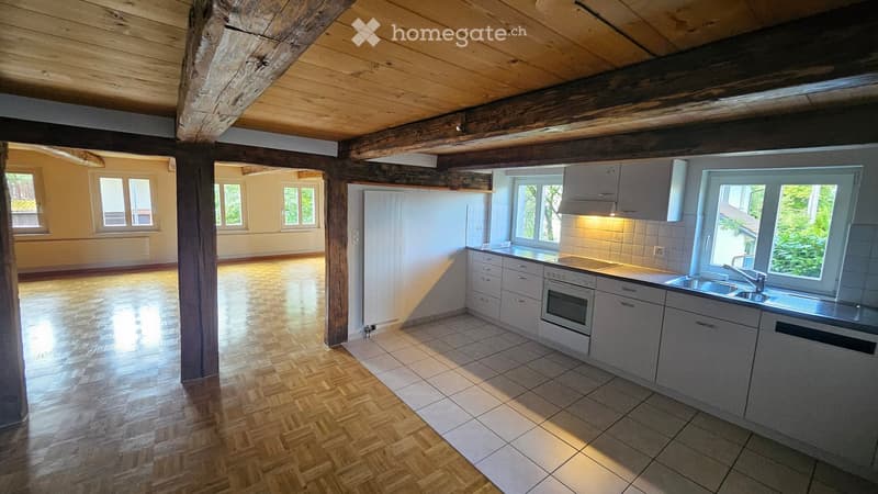 Modern kitchen with light grey cabinets, stainless steel sink, and oven. Wooden herringbone floor and exposed wooden beams on the ceiling.