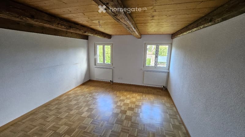 Empty room with wooden flooring, white walls, exposed wooden beams on the ceiling, and two windows with white frames. Radiators are visible beneath the windows.