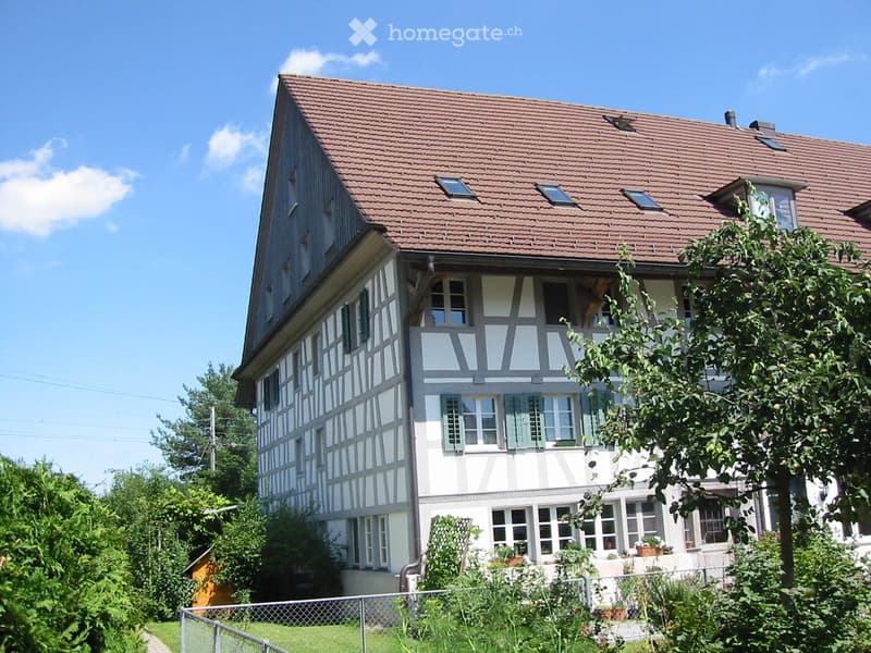 Two-story house with a red-tiled roof, white and grey facade, multiple windows, and a fenced garden area.