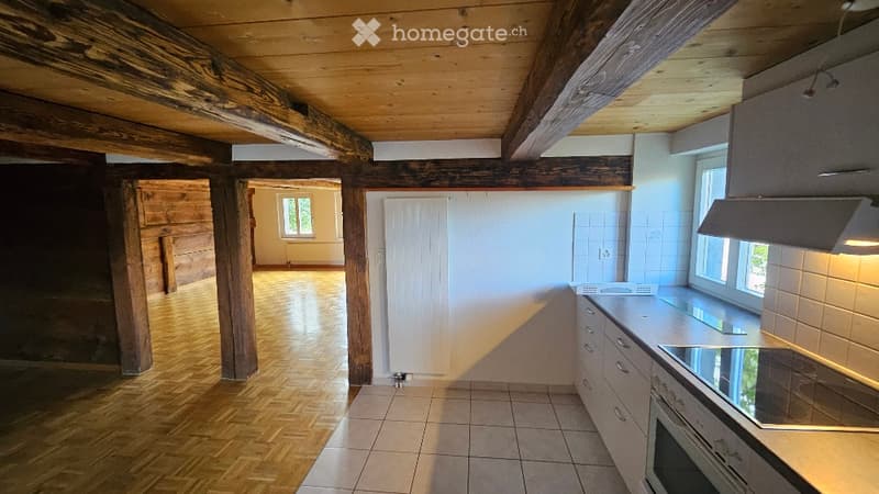 Bright kitchen with wooden beams, white tiled walls, and modern appliances including a stove and oven. The space features wooden flooring and ample natural light from large windows.