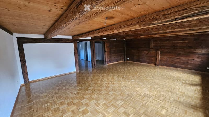Empty room with wooden floor and exposed wooden beams on the ceiling. The walls are a mix of light plaster and dark wood paneling. There are no visible furnishings or decorations.