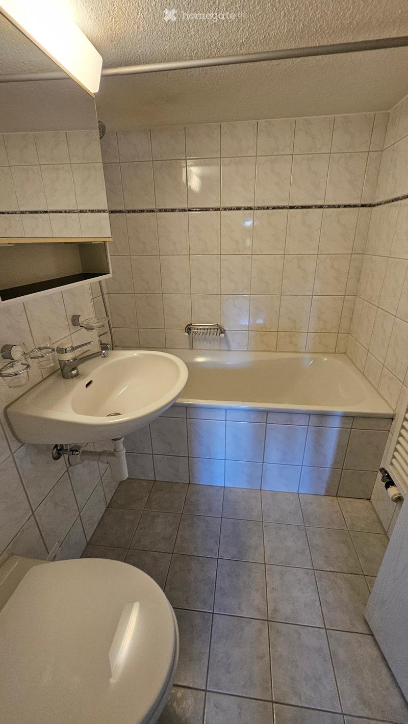 Bathroom with a sink, bathtub, toilet, and radiator. The walls are tiled with beige tiles and the floor is tiled with grey tiles.