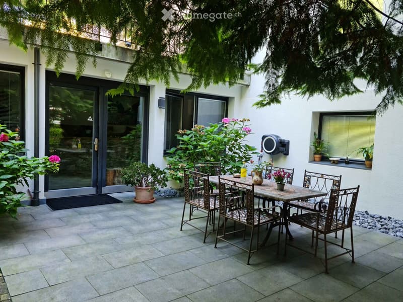 A well-maintained patio area with a tiled floor, a metal table surrounded by chairs, potted plants, and a vase on the table. The patio is enclosed by a modern building with large glass doors and windows, and is shaded by trees.