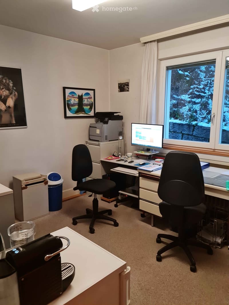 Office space with two desks, two chairs, a computer screen, a printer, a coffee machine, and framed pictures on the wall.