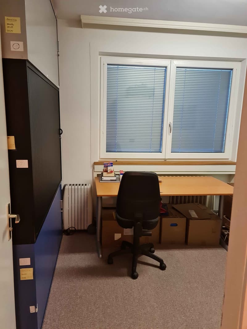 A small, furnished room with a desk, chair, radiator, and several cardboard boxes. The room has a window with blinds and a door with a blue panel.