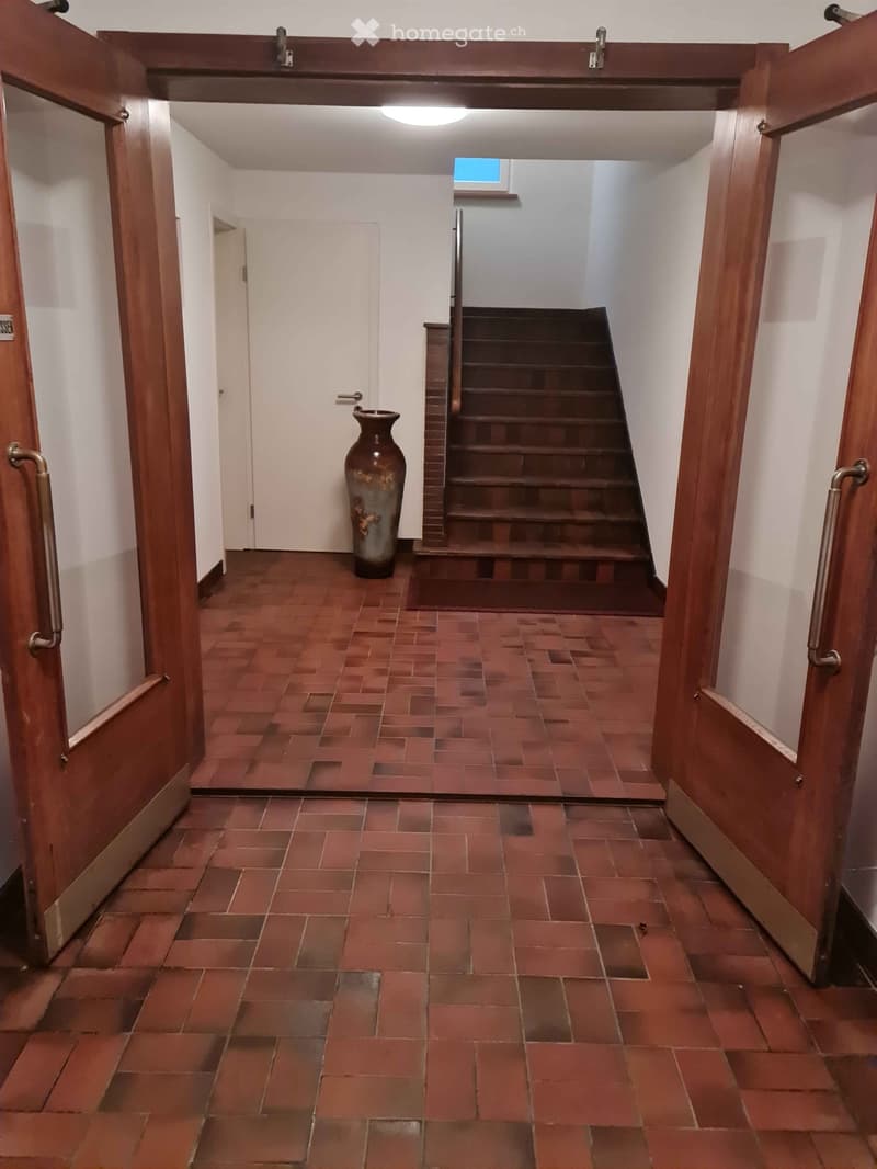 Hallway with red brick tiles, wooden doors, staircase leading upwards, and a decorative vase in the center.