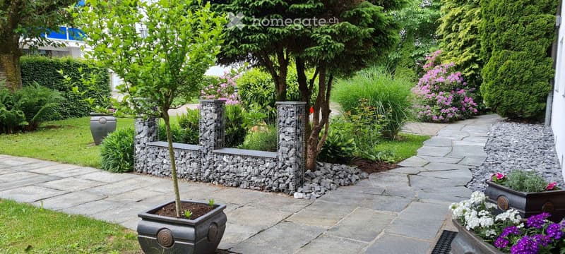 Well-maintained garden with stone pathways, potted plants, shrubs, and a small tree. Features decorative stone walls and a variety of flowering plants.