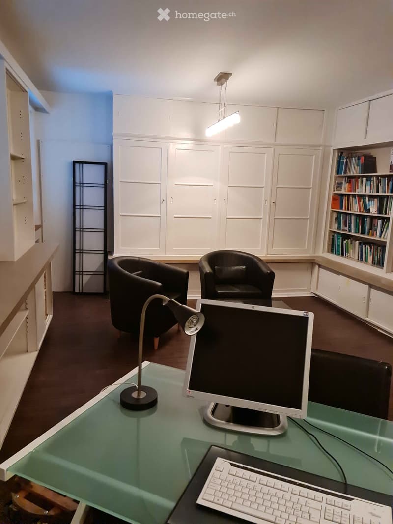 Modern office space with a glass desk, laptop, keyboard, table lamp, two black armchairs, and built-in white cabinets with a bookshelf filled with books.