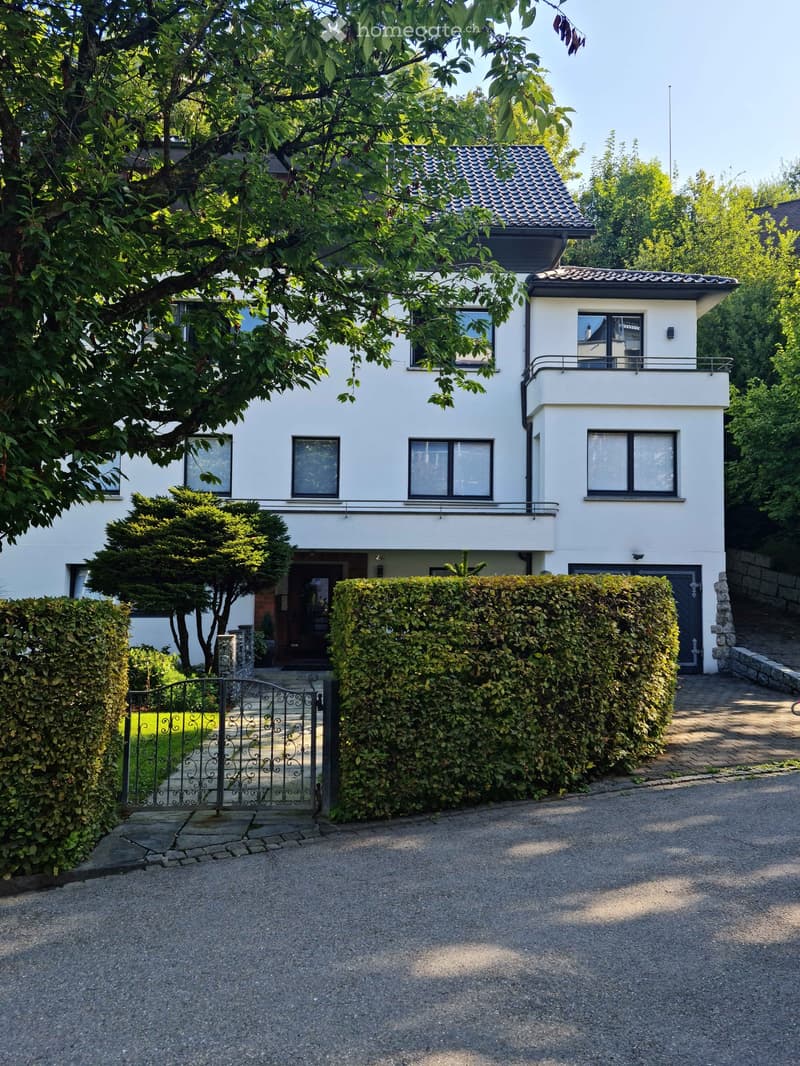 Two-story white house with a tiled roof, multiple windows, and a small balcony on the upper level. The house is surrounded by greenery, including trees and shrubs, with a gated entrance and a driveway leading to the house.