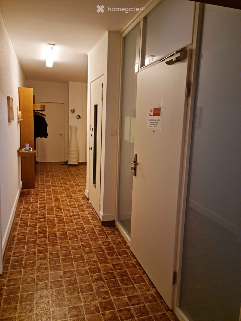 A narrow hallway with tiled flooring, white walls, and several doors. A small wooden shelf with a vase and a coat is visible on the left side.