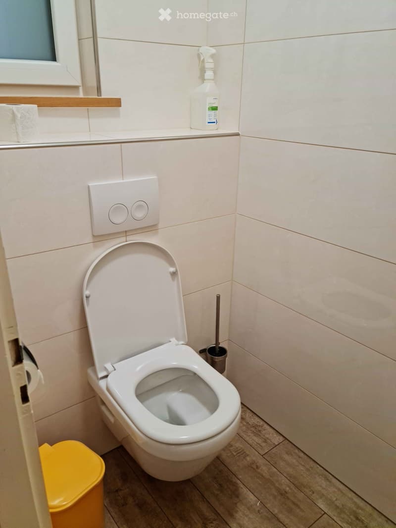 Modern bathroom with a white toilet, toilet paper on a holder, a toilet brush in a holder, a cleaning bottle on the shelf, and a yellow trash can on the floor.