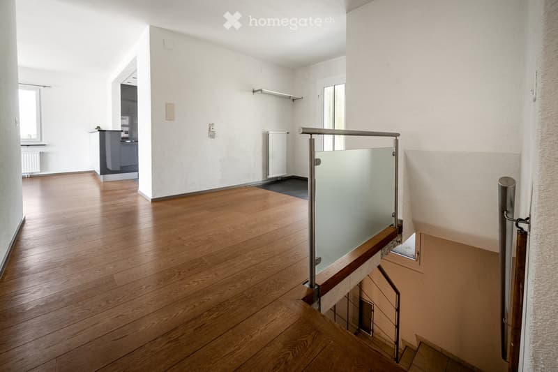 Empty hallway with wooden flooring, white walls, and a modern glass railing leading to an adjacent room.