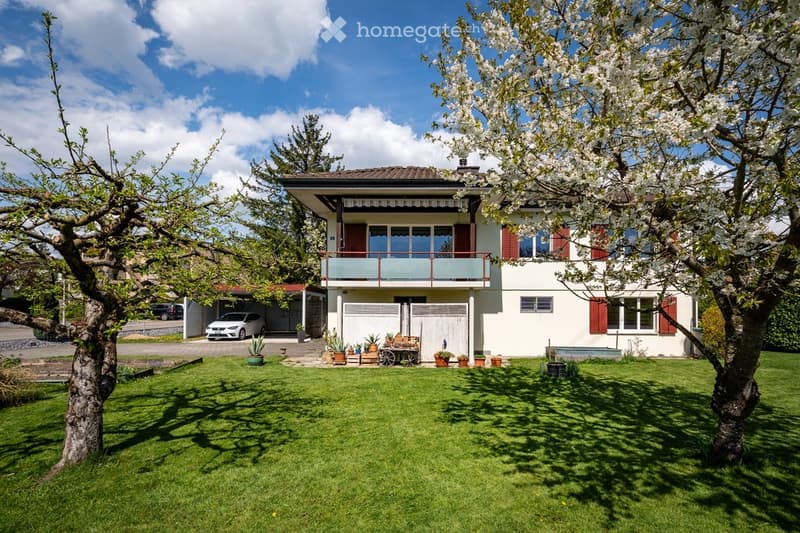 Two-story house with white exterior, red shutters, and a balcony on the upper floor. The house features a well-maintained lawn, several potted plants, and a small garden area. A garage and parking space are visible to the left.