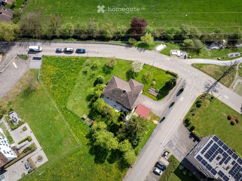 Aerial view of a residential area with a two-story house, surrounded by green lawns, trees, and a parking area with several cars.