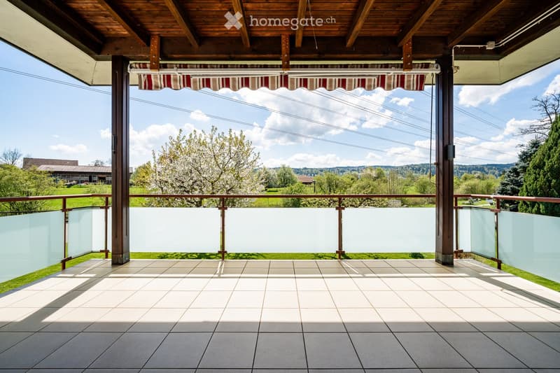 Balcony with tiled floor, wooden beams, glass railing, and striped awning, overlooking a green landscape with trees and hills.