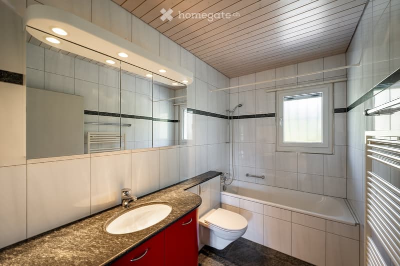 Modern bathroom with a large mirror, granite countertop, sink, bathtub, toilet, and radiator. Features a wooden ceiling and tiled walls.