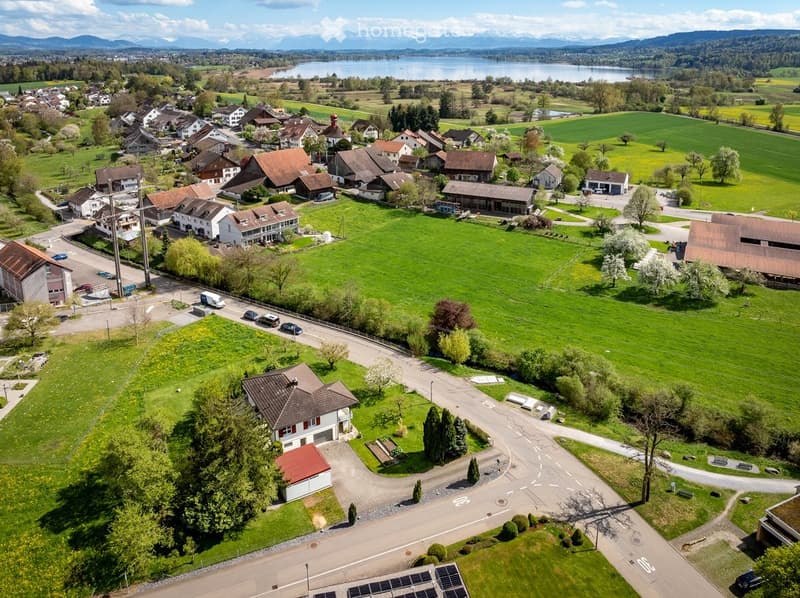 Aerial view of a small village with houses, green fields, and a lake in the background.