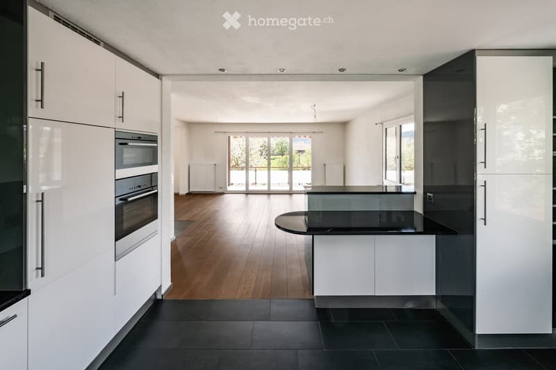 Modern kitchen with white cabinets, stainless steel appliances, black countertops, and a central island. The space features wooden flooring in the adjacent living area and large windows providing natural light.