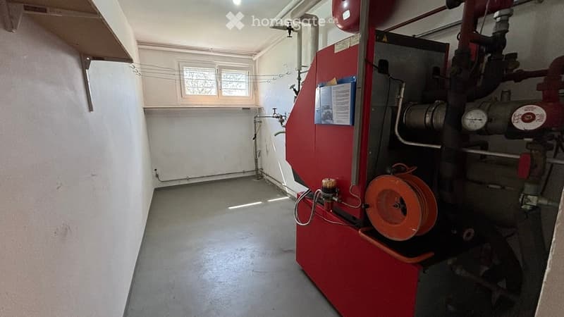 Utility room with a heating boiler, pipes, gauges, and a small window. The room is empty with no furniture.