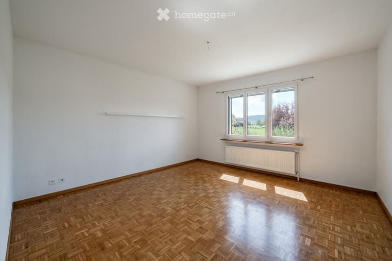 Empty room with wooden parquet flooring, white walls, and large windows offering a view of greenery outside. The room features a radiator under the windows and a small shelf on one wall.