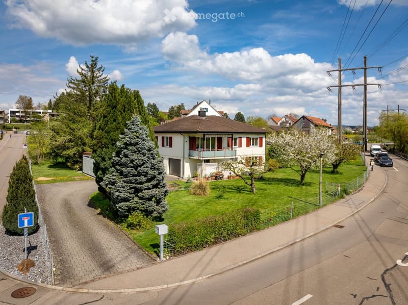 Two-story house with white exterior, red shutters, and a balcony. The house is surrounded by a well-maintained lawn, trees, and a driveway leading to a garage. There are several parked cars on the street.