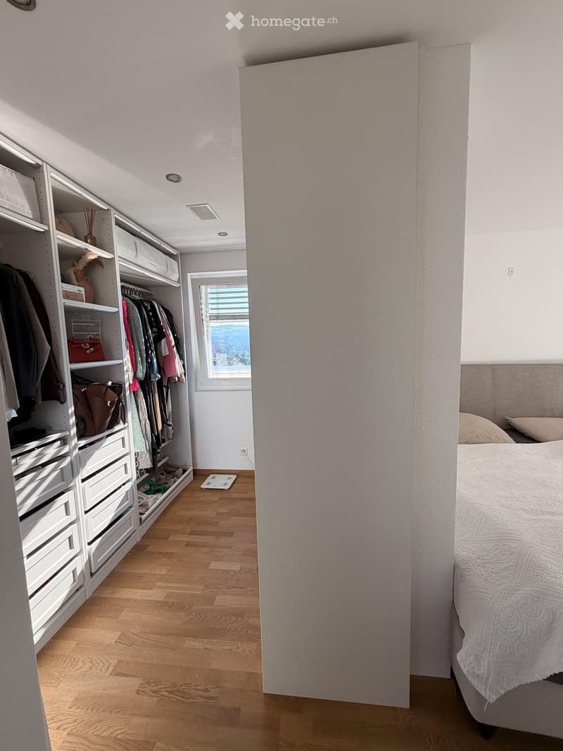 A modern bedroom with a large bed, white wardrobe with shelves and drawers, and a window offering a scenic view.