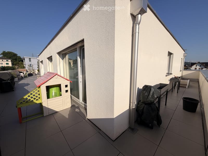 Balcony with tiled floor, white walls, and a small playhouse with a red roof. There is a covered grill, a covered chair, and a table with chairs.