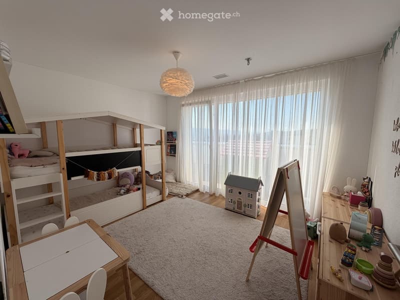 Brightly lit children's room with bunk beds, a small table, a toy house, and various toys scattered around. The room features large windows with white curtains and a cozy rug on the floor.
