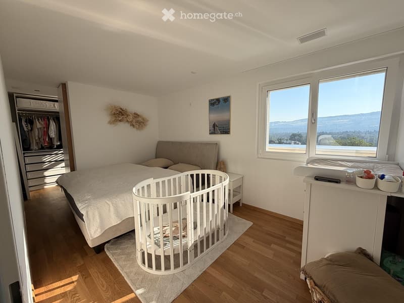 Bright bedroom with large windows offering a scenic view, featuring a bed, nightstand, cradle, wardrobe, and decorative items.