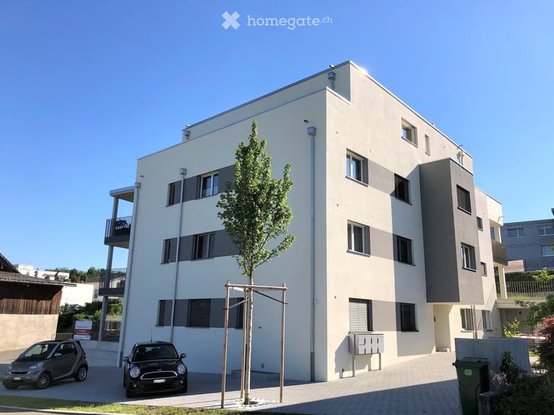Modern apartment building, white exterior, multiple windows, balconies, parking area, small tree in front