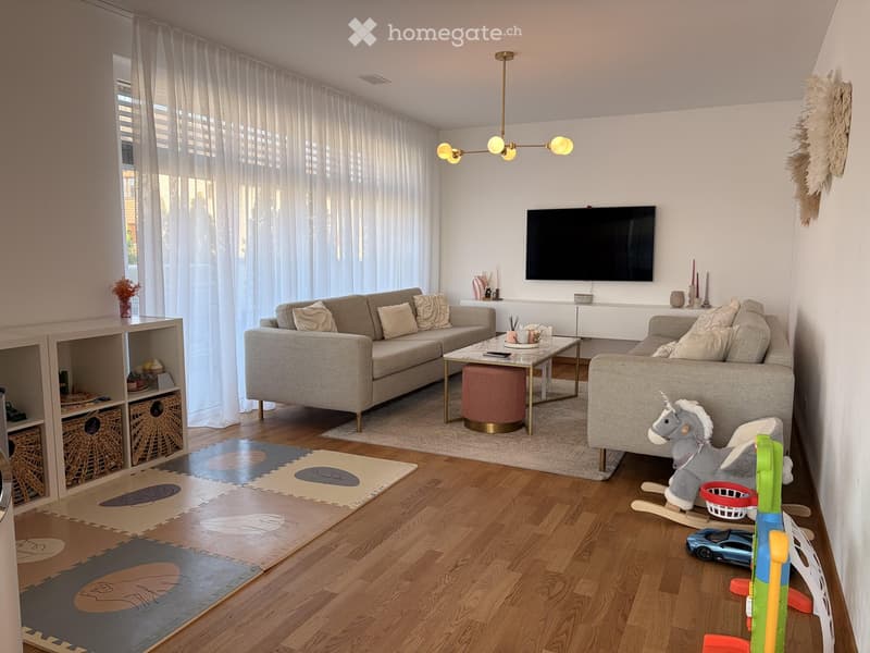 Modern living room with hardwood floors, large windows with sheer curtains, a sectional sofa, coffee table with ottoman, wall-mounted TV, and various toys including a rocking horse and toy car.