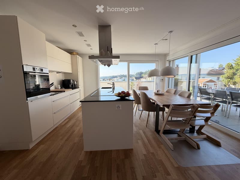 Modern kitchen with white cabinets, a central island, and a dining area. Features include an oven, a coffee machine, and a large window with a view of the outdoor area.
