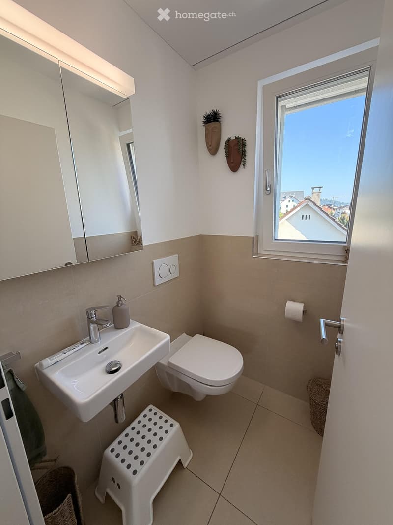 Modern bathroom with a white sink, chrome faucet, toilet, mirror cabinet, and a small stool with black polka dots. The room features beige tiles and a window offering a view of the outside.