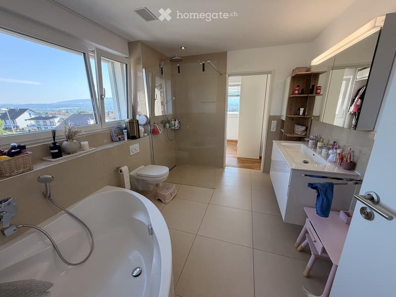 Modern bathroom with a bathtub, toilet, sink, mirror, and shower. Features large windows with scenic views, built-in shelves, and decorative items.
