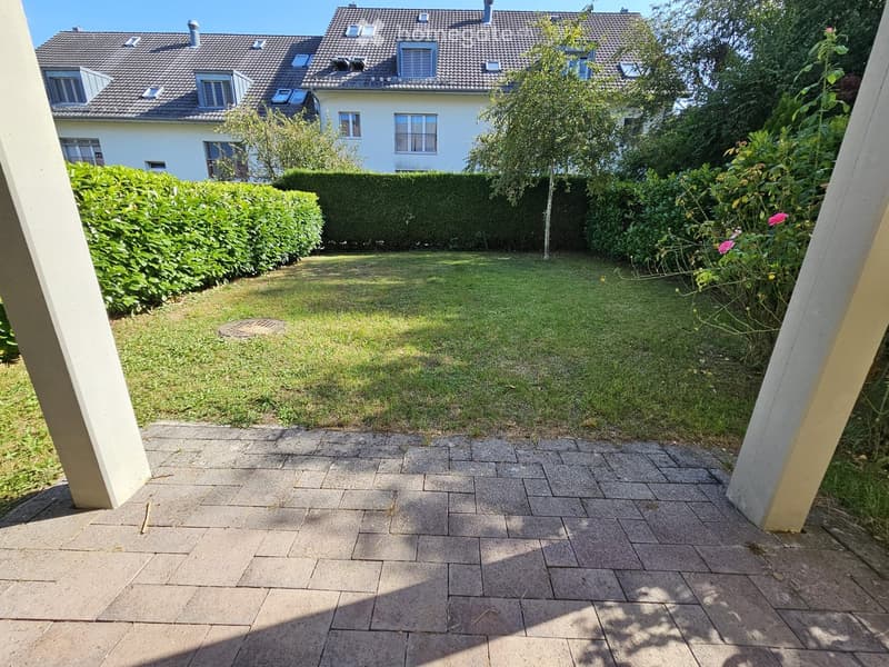 Garden area with a well-maintained lawn, trimmed hedges, and a paved patio area with stone tiles. A house with multiple windows and a tiled roof is visible in the background.