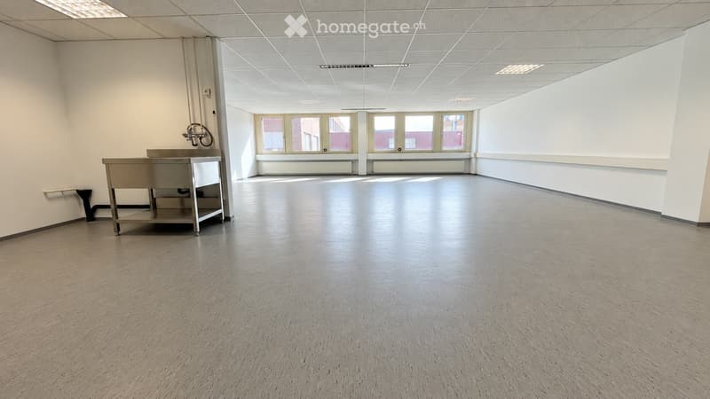Empty room with bare walls, a grey floor, and a small metal utility table with a sink in the corner. Windows at the far end provide natural light.