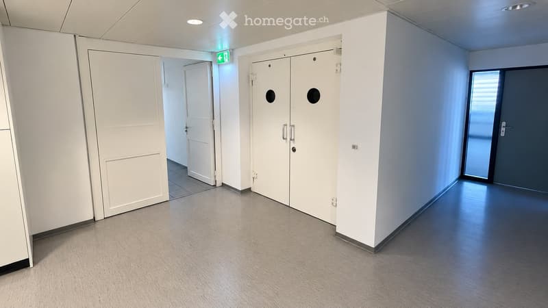 Clean, modern hallway with white walls, grey carpeted floor, white doors, and an emergency exit sign above a door.