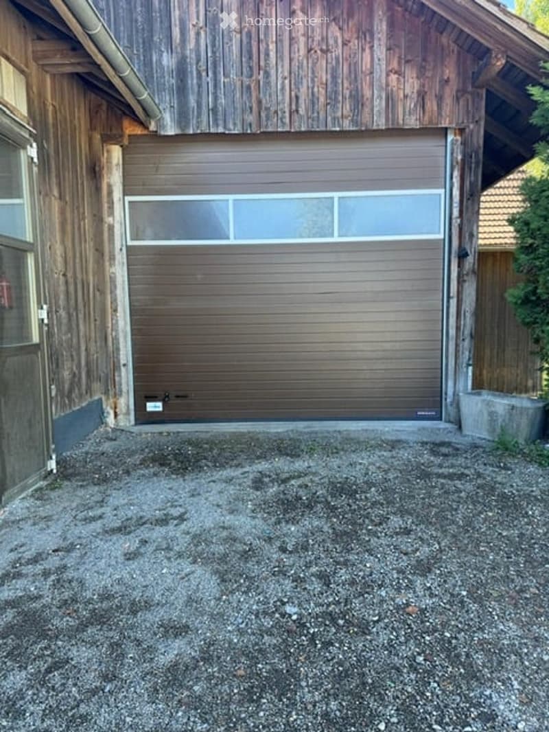 Wooden garage with a large, closed garage door and small windows above it. The surrounding area is gravel-covered.