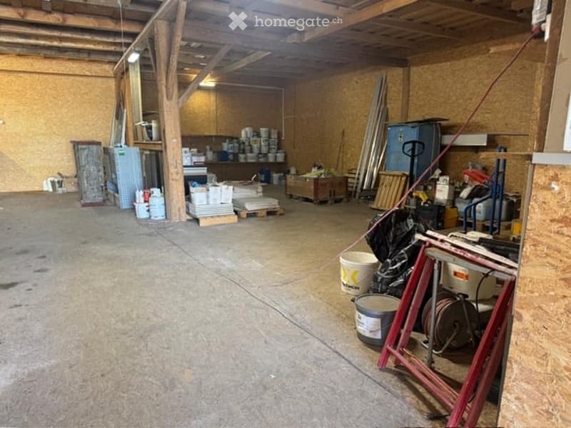 Empty warehouse or garage space with exposed wooden beams, concrete floor, and scattered construction materials and tools.