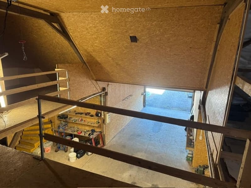 An attic space with exposed wooden beams, a metal staircase leading up, and a workbench with tools and storage items. The area is unfinished with visible insulation and a concrete floor.