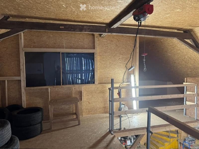 An attic space with exposed wooden beams, unfinished walls, and a single window. There are a few tires, a wooden stool, and a small staircase leading down.