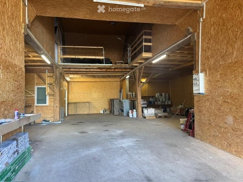 Empty warehouse or industrial space with exposed wooden beams, concrete floor, and minimal furnishings. Stairs lead to an upper loft area.
