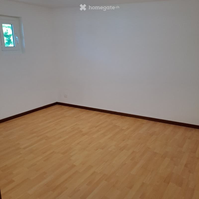 Empty room with light wooden flooring and white walls, featuring a small window with a view of greenery.
