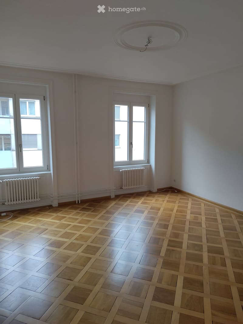 Empty room with wooden parquet flooring, white walls, large windows, and radiators. No furniture or equipment present.