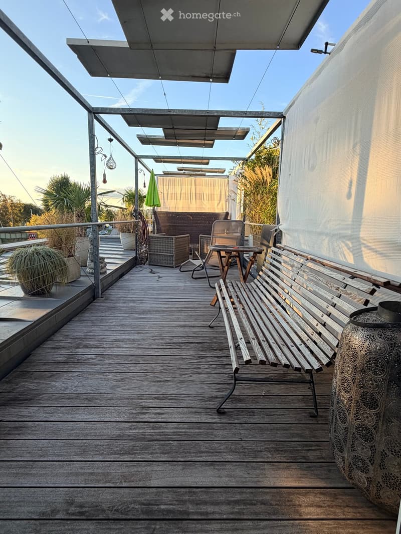 Wooden deck with a bench, small table, chairs, wicker sofa, and an umbrella, surrounded by plants and a railing.